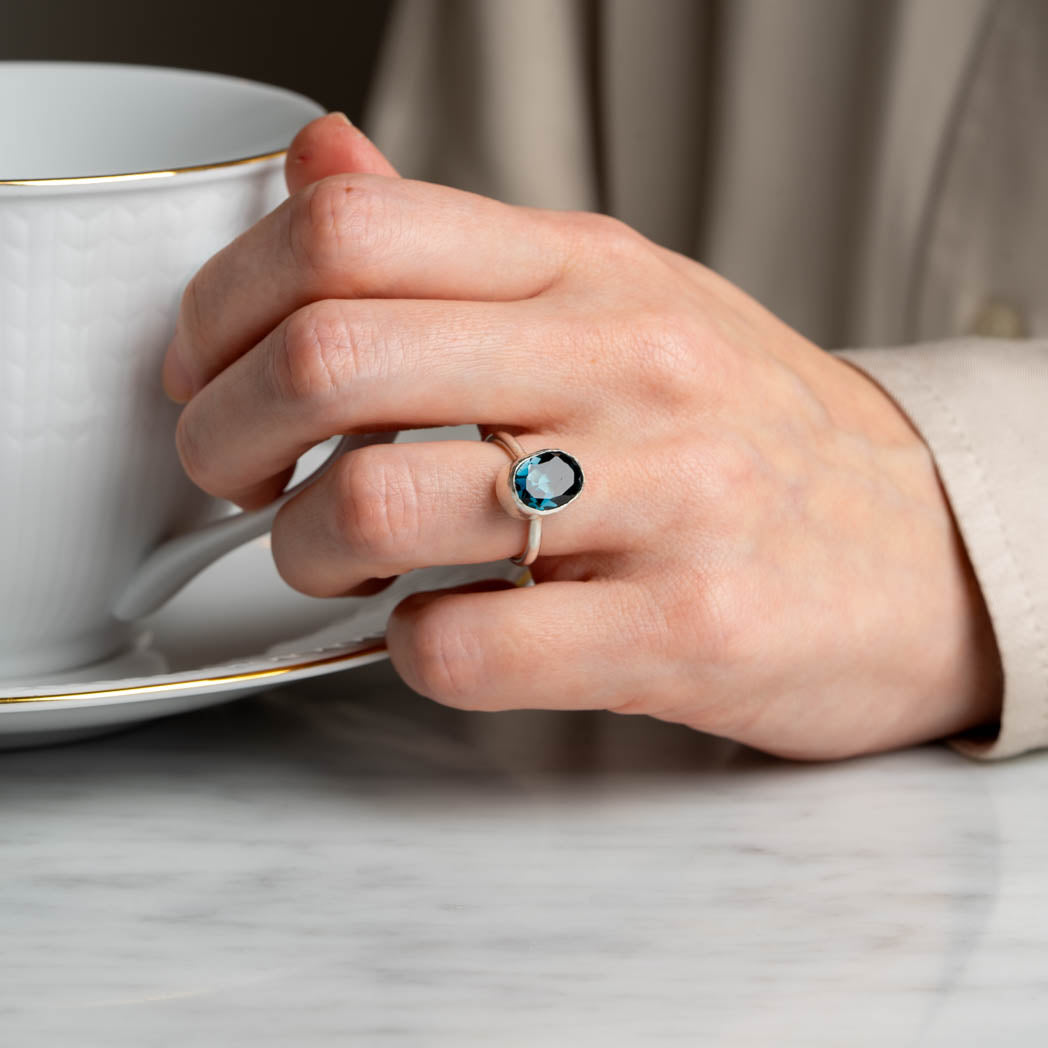 Lifestyle-bild av en hand med vår London Blue Topas-ring i äkta silver, fasettslipad sten, vilande på ett bord och hållande en kopp.