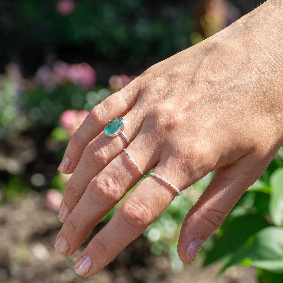 Kvinnas hand med vår handgjorda silverring, prydd med en rosenslipad smaragd, vackert kontrasterande mot en rabatt full av blommor.